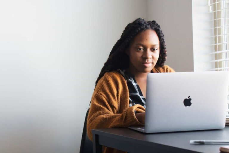 A parent with braided hair and wearing a brown sweater sits at a desk, looking at an open laptop. Light from a window illuminates the scene as they research websites for special needs resources.
