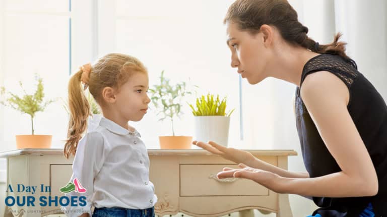 An adult woman kneels and talks seriously to a young girl, emphasizing positive discipline, in a bright room with plants and a desk. Logo text reads, “a day in our shoes. ”.