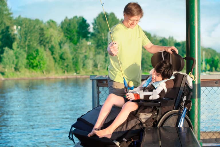 An adult helps a child in a wheelchair hold a fishing rod on a lakeside dock, highlighting fishing programs that create opportunities for kids with disabilities to enjoy the outdoors among trees and water.