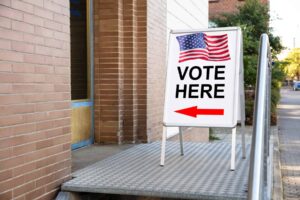 A sign with the american flag reads "vote here" with a red arrow pointing left, placed on an accessible ramp outside a brick building entrance, highlighting available election resources for individuals with disabilities.
