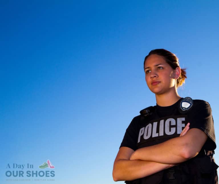 A police officer, embodying community support, stands with arms crossed against a clear blue sky. Clad in a black shirt emblazoned with "police" and a utility vest, he represents the teamwork akin to iep parent participation. The logo text "a day in our shoes" rests at the bottom left.