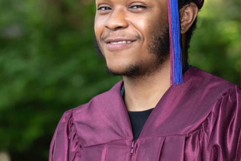 A man walking at graduation, but not graduating, wearing a graduation gown and smiling.
