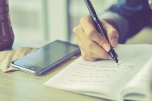 A person is writing with a pen in an open notebook, likely taking detailed notes. A smartphone, perhaps used to record the iep meeting, is placed on the table beside the notebook.