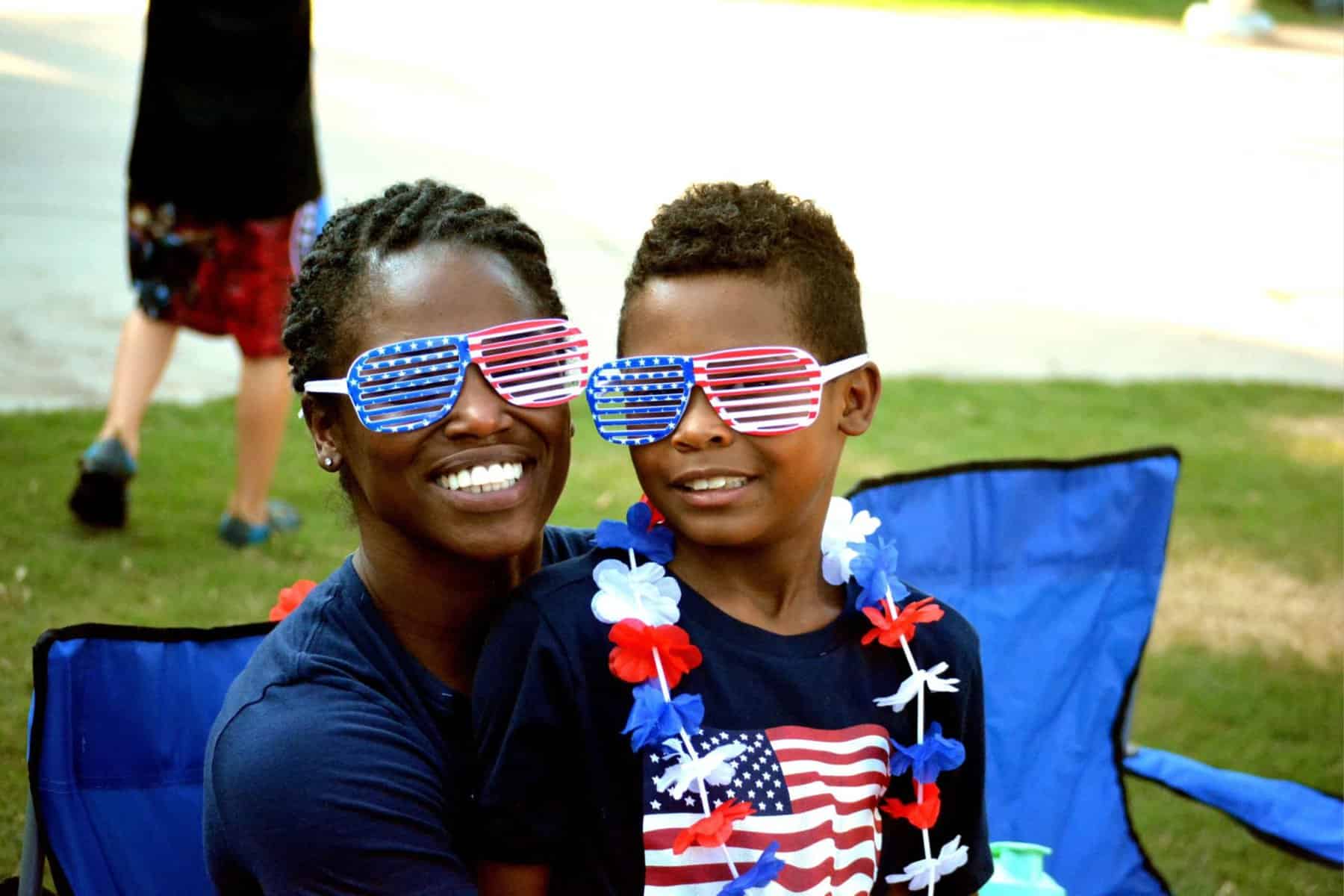 A woman and a young boy wearing sunglasses with american flag designs, smiling at a festive outdoor event, embodying patriotism for kids.