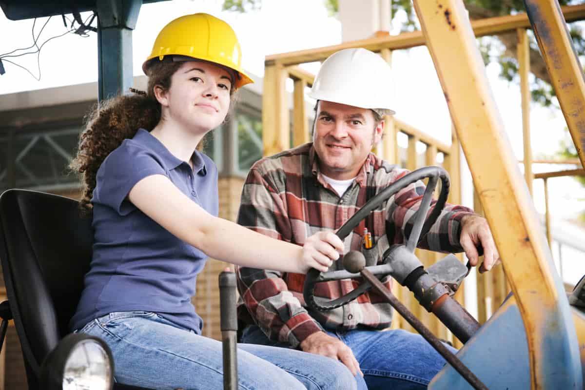 Two people wearing hard hats are sitting in a construction vehicle. One person, seated at the wheel, looks ahead, while the other, standing beside, smiles at the camera—showcasing real-world skills from iep vocational assessments on their path to success.