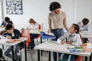 A teacher holding a notebook provides specially designed instruction to a student at her desk in a classroom while other students are focused on their work.