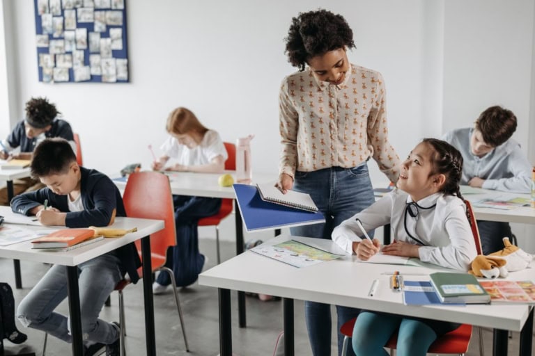 A teacher holding a notebook provides specially designed instruction to a student at her desk in a classroom while other students are focused on their work.