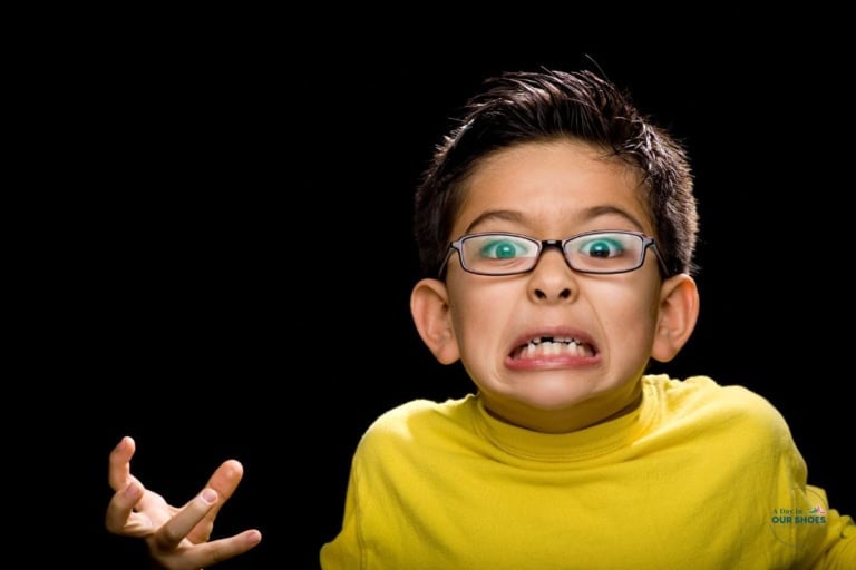 A young boy wearing glasses and a yellow shirt grimaces and raises one hand, standing against a black background—an expression that may reflect moments of pda or autism masking.