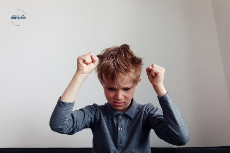 Young boy with brown hair in a grey shirt raises both fists and frowns, appearing frustrated or upset—a moment that may reflect autism masking or the challenges of pda—against a plain white background.