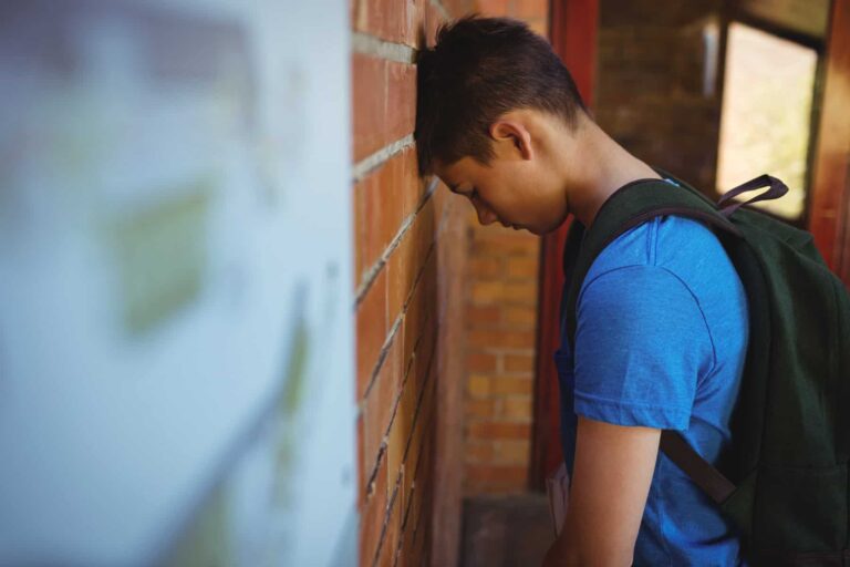 A young person in a blue shirt, who happens to have an iep but is still struggling academically, rests their forehead against a brick wall while wearing a backpack.