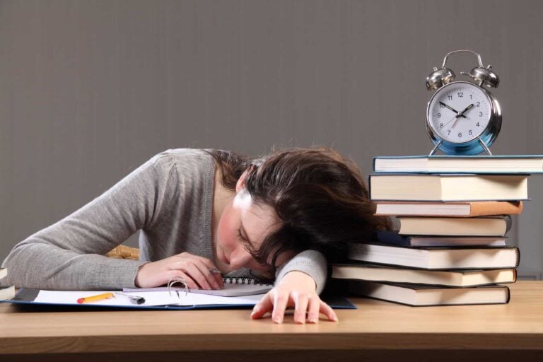 A woman utilizing executive function skills by sleeping on a desk with books and an alarm clock nearby.