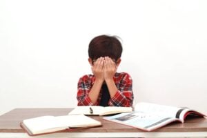 A boy is sitting at a desk, feeling overwhelmed with work completion tasks. He reluctantly covers his face with his hands, struggling to focus on the preferred and non-preferred tasks assigned to him as per