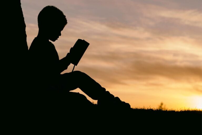 A silhouette of a boy reading a book at sunset, working to improve his reading comprehension.