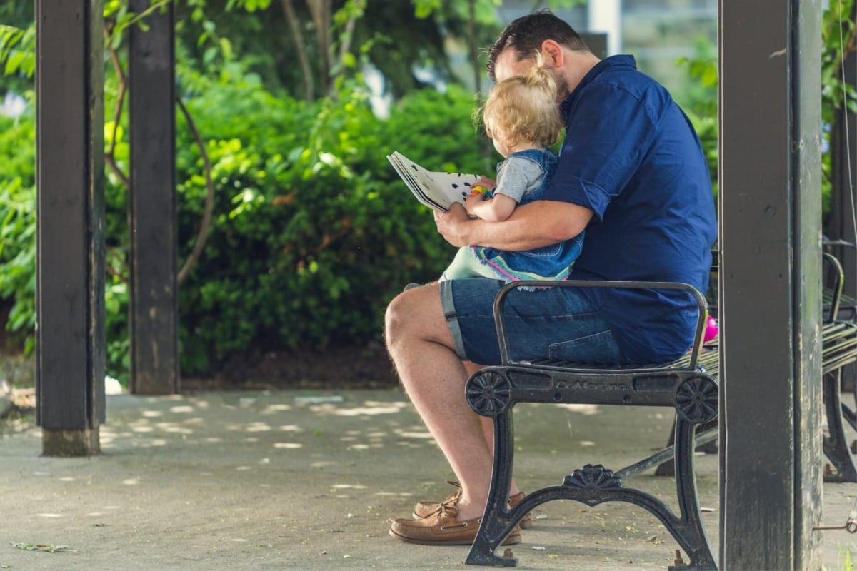 A man improving reading comprehension by fluently reading a book to his child on a bench.