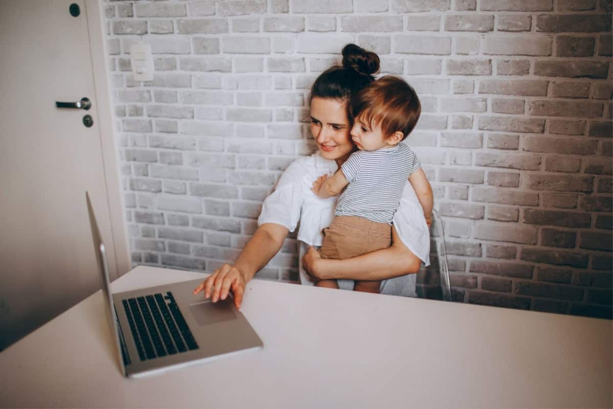 A stay-at-home mom multitasking by holding a baby while working on a laptop.