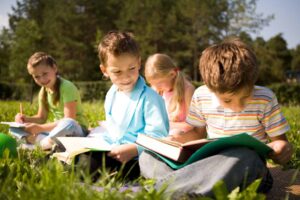 A group of children participating in an extended school year sitting in the grass reading books.
