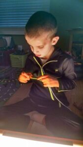 A young boy in a tracksuit sits cross-legged on the floor, focused on holding a green object under soft lighting. Designed with iep goals for low functioning students in mind, the room features a toy basket and a mat with road patterns to foster engagement and learning.