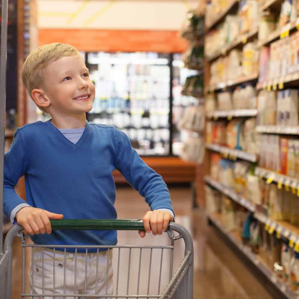 A young boy with a smile participates in community-based instruction, holding onto a shopping cart in a grocery store aisle.