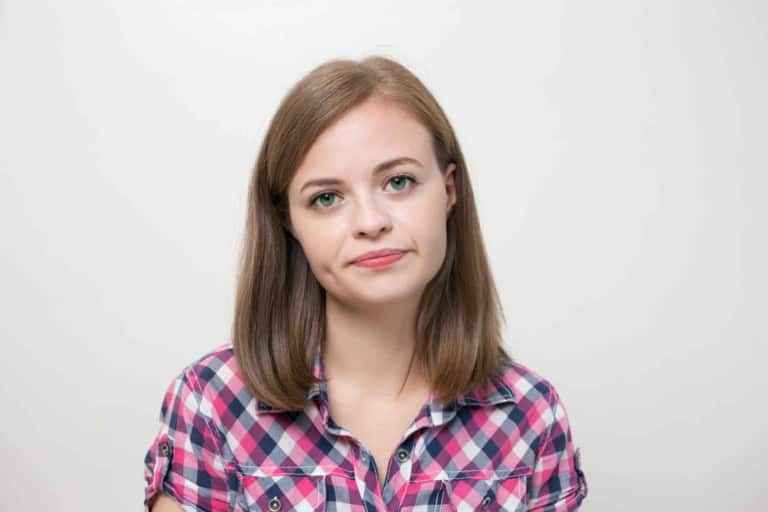 A woman with shoulder-length brown hair wearing a pink and blue plaid shirt, looking at the camera w.