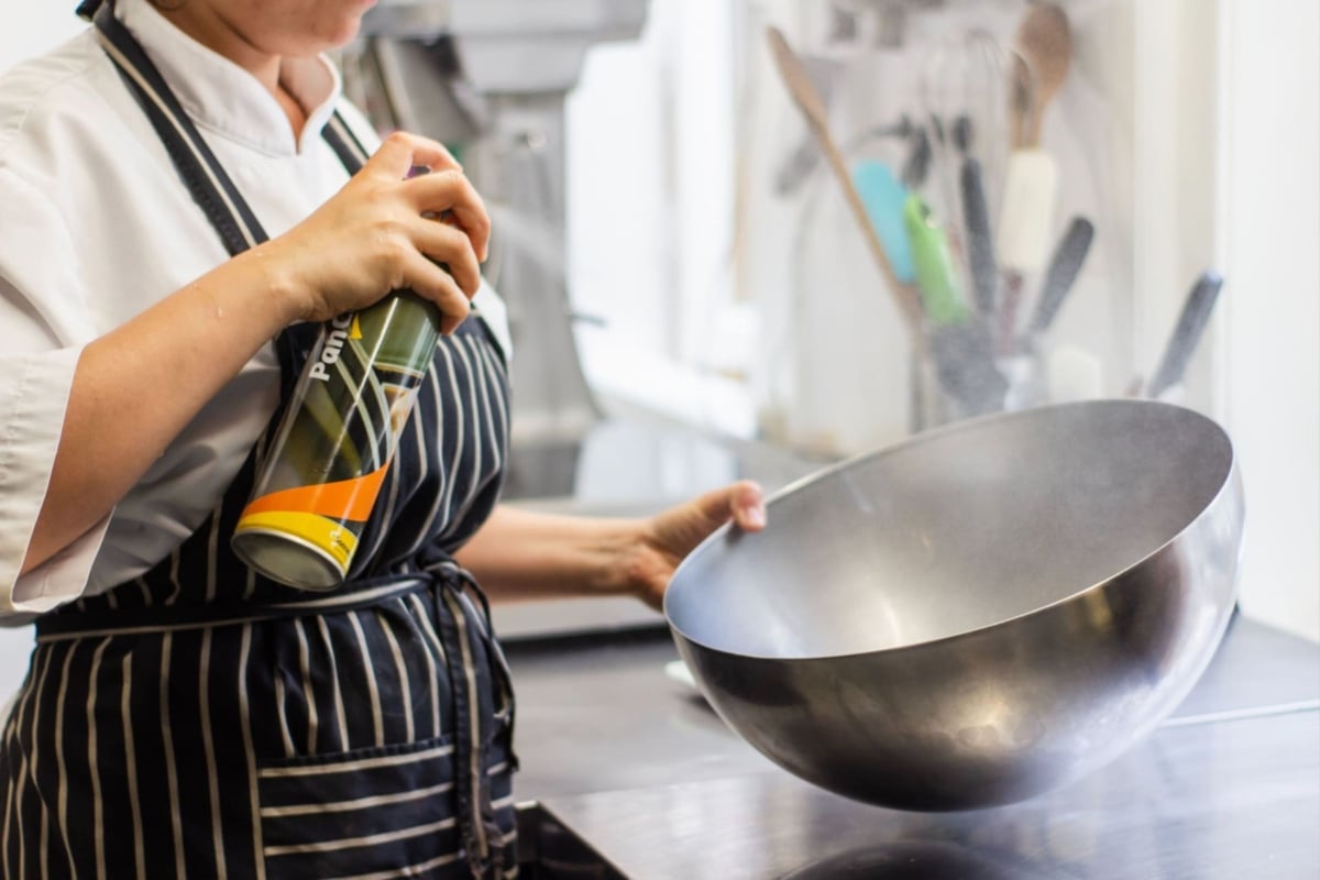 A chef in a striped apron is holding a large metal wok in one hand and a bottle of oil in the other, preparing to pour the oil into the wok as part of vocational i
