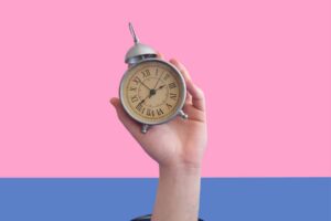 A hand is holding an analog alarm clock against a split pink and blue background, demonstrating telling time iep goals.