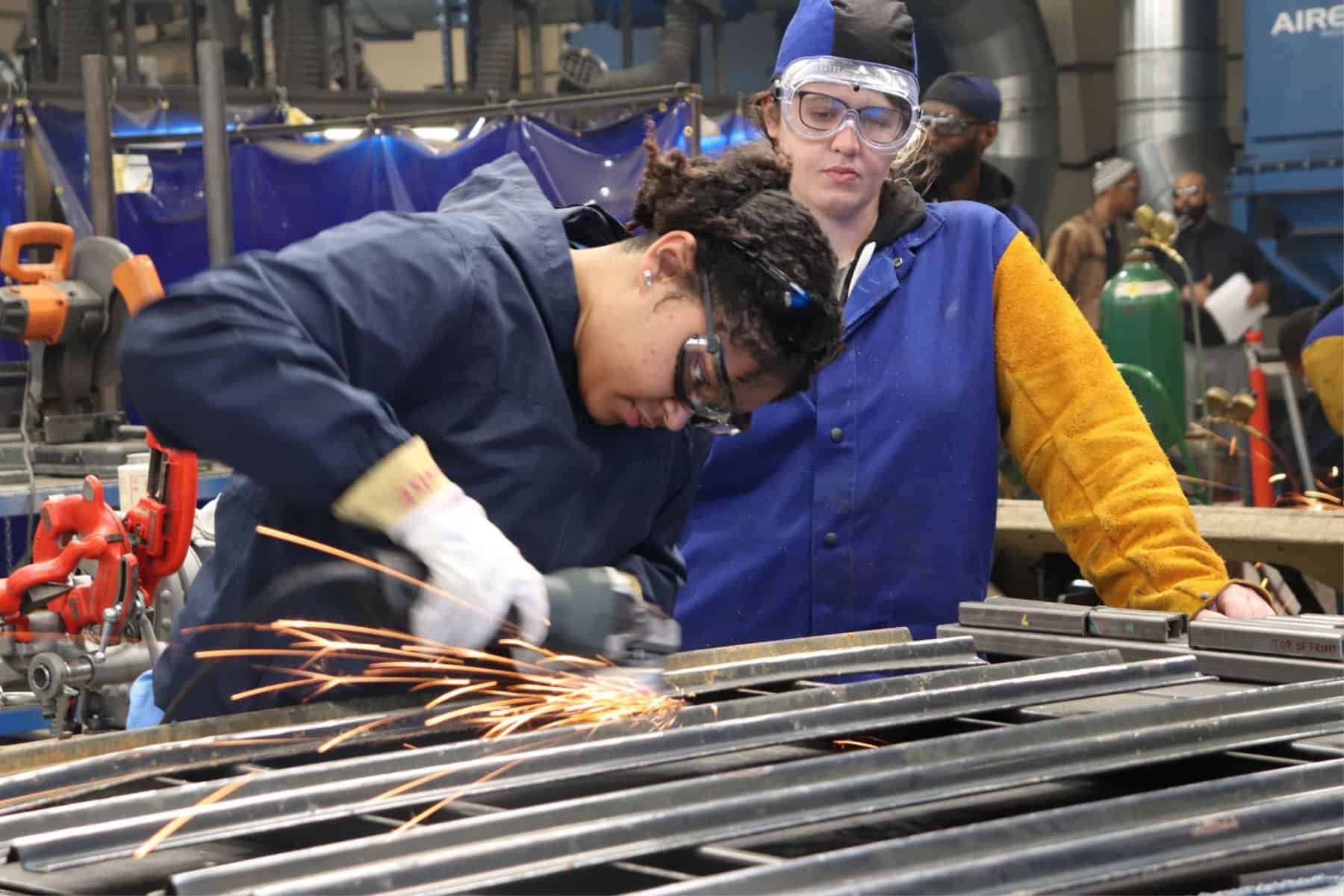 A worker in protective gear uses an angle grinder on a metal piece to achieve vocational iep goals while another person observes in the background of an industrial setting.