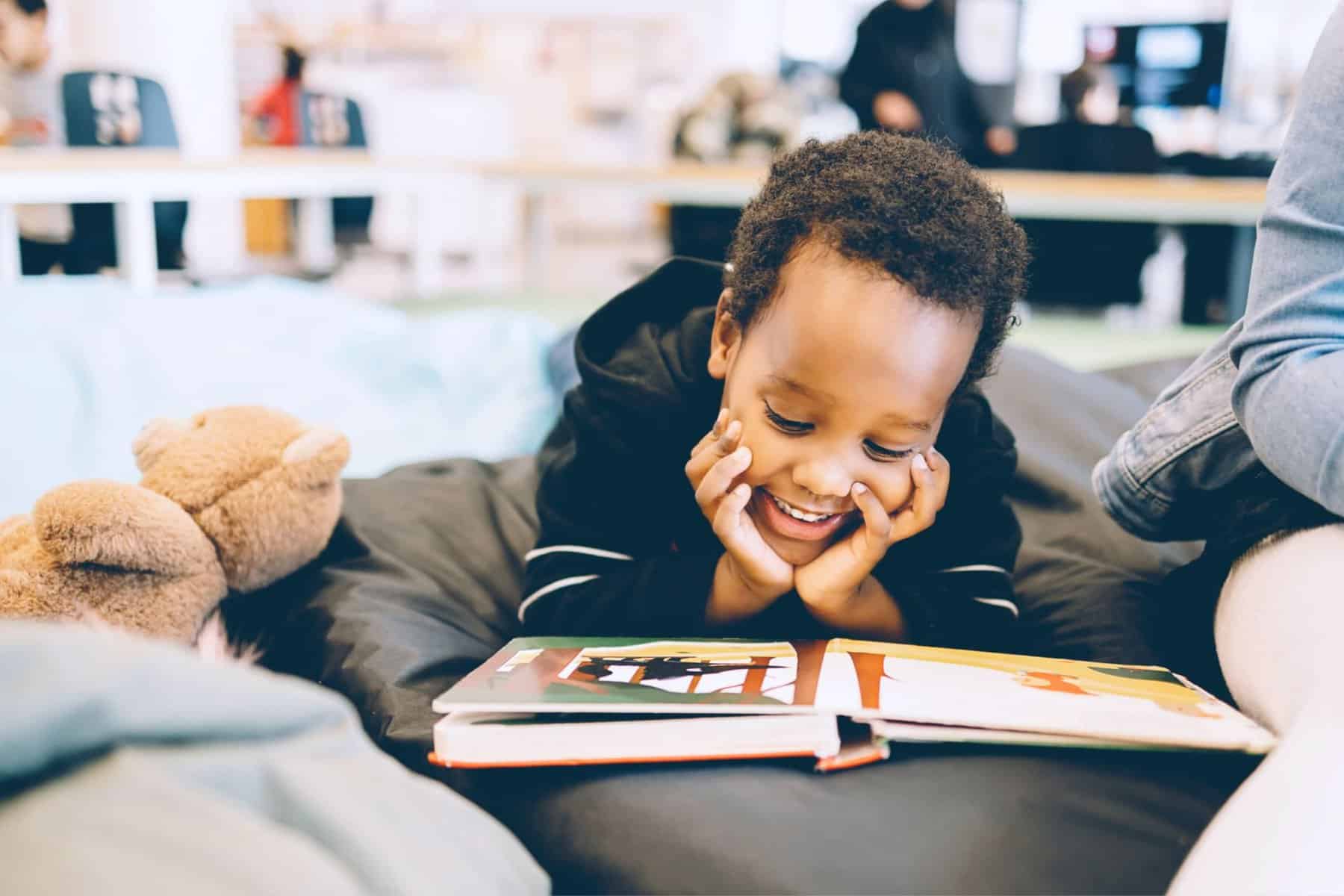 A young child lying on their stomach on a cushioned surface, smiling while looking at an open picture book to improve basic reading skills, with a plush toy nearby.