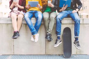 Four teenagers sitting on a ledge, engrossed in their devices—a tablet, a book, and smartphones—beside a skateboard. They effortlessly blend technology with fun, almost like they're in their own social skills game for teenagers.