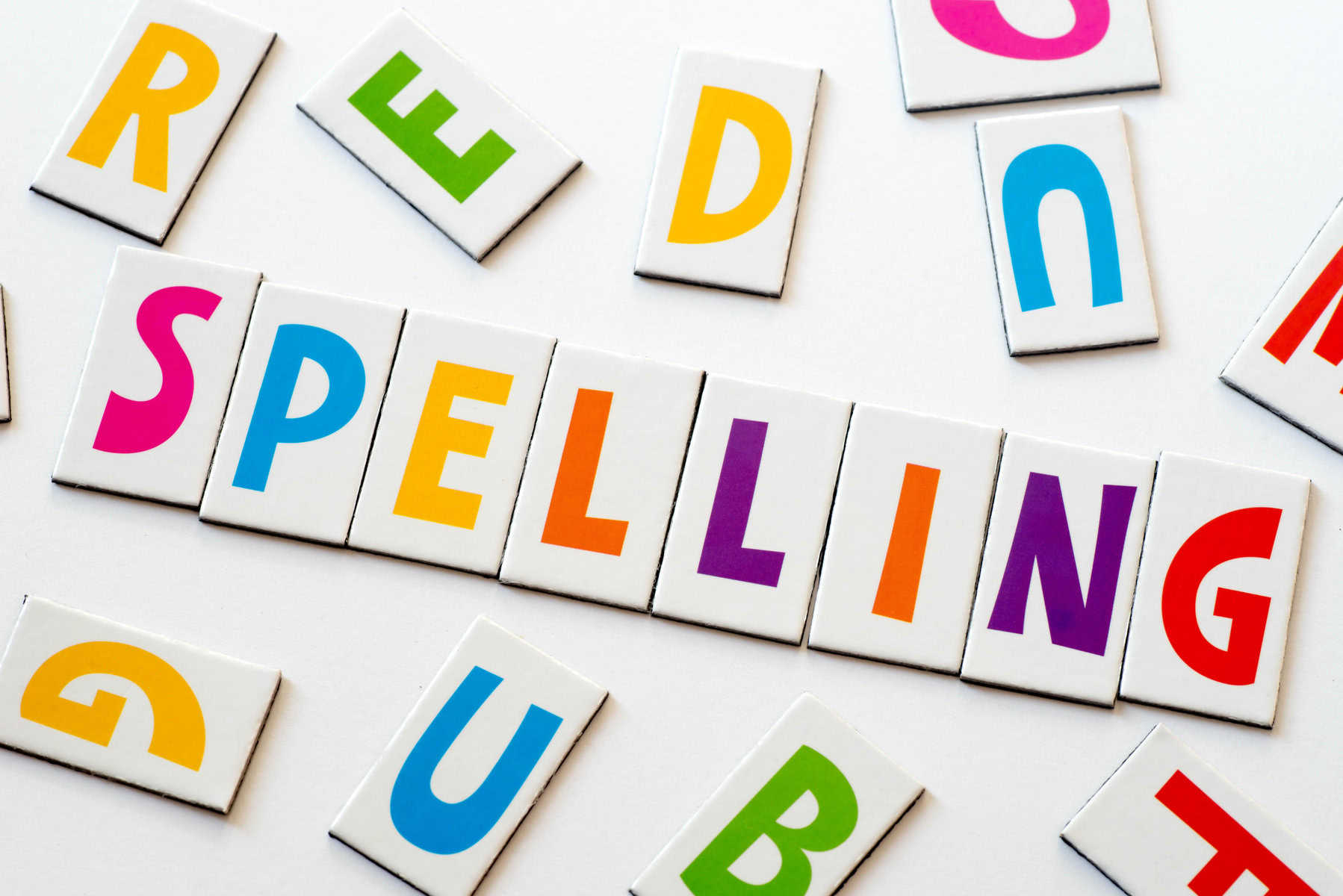Colorful letter tiles spell "spelling" on a white surface, surrounded by other scattered tiles. These playful tools can be instrumental in achieving spelling iep goals, making learning fun and visually engaging.