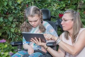 In a lush garden with vibrant green foliage, a woman is training a girl in a wheelchair on using aac apps on a tablet.