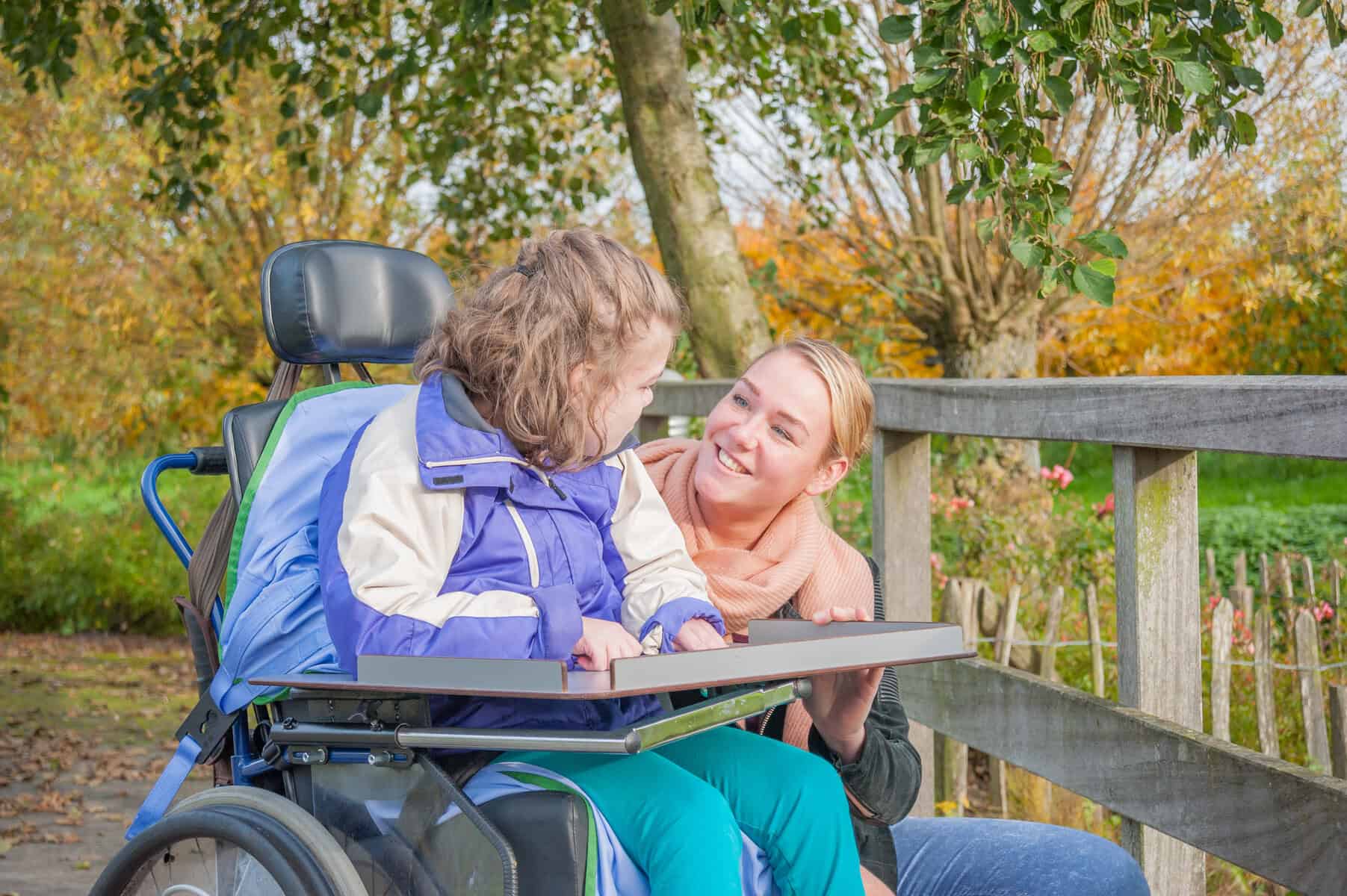 A woman in a wheelchair supporting students with severe and profound needs using a communication device smiles at another woman leaning close by a park bench.