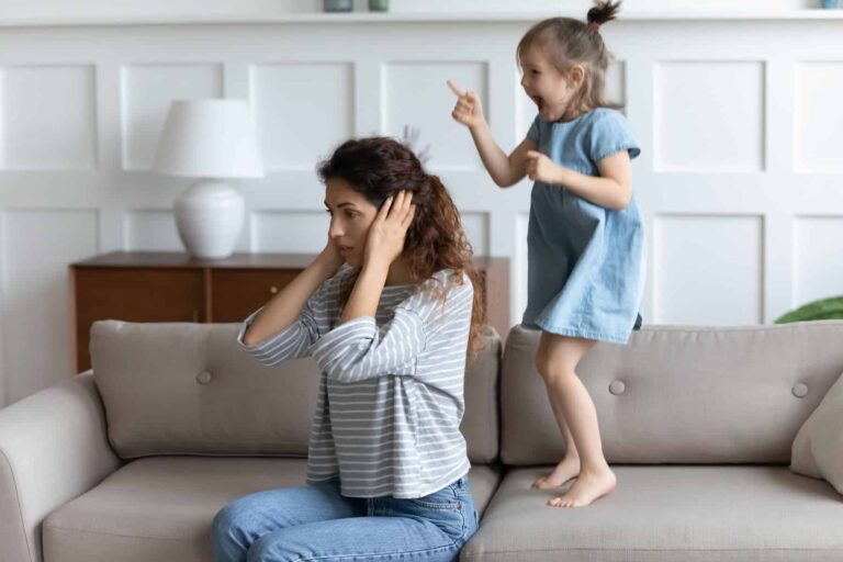 A frustrated mother sitting on a couch practices active ignoring as she covers her ears while her young daughter, standing on the couch, points and appears to be shouting.