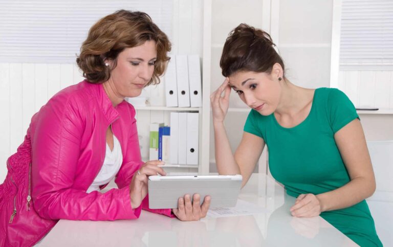 Two women are seated at a table, looking at a tablet. One woman in a pink jacket holds the device, discussing ai for iep writing, while the other in a green shirt appears concerned.
