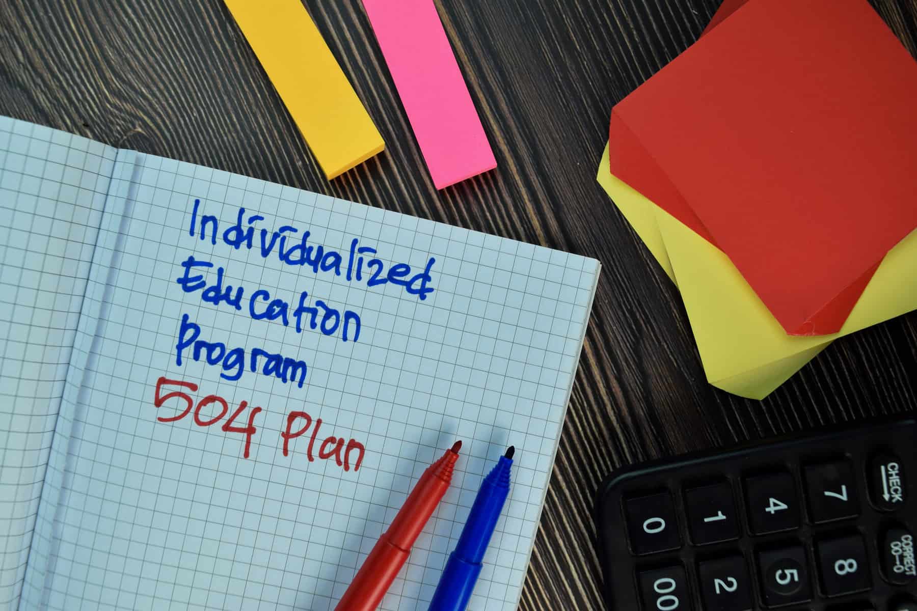 Notebook with handwritten text "individualized education program 504 plan" in blue and red ink, two pens, sticky notes, and a calculator on a desk. A small note asks, "does a 504 plan expire?