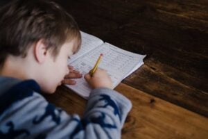 A young boy in a blue shirt develops his test-taking skills by writing in a workbook with a yellow pencil, sitting at a wooden table.