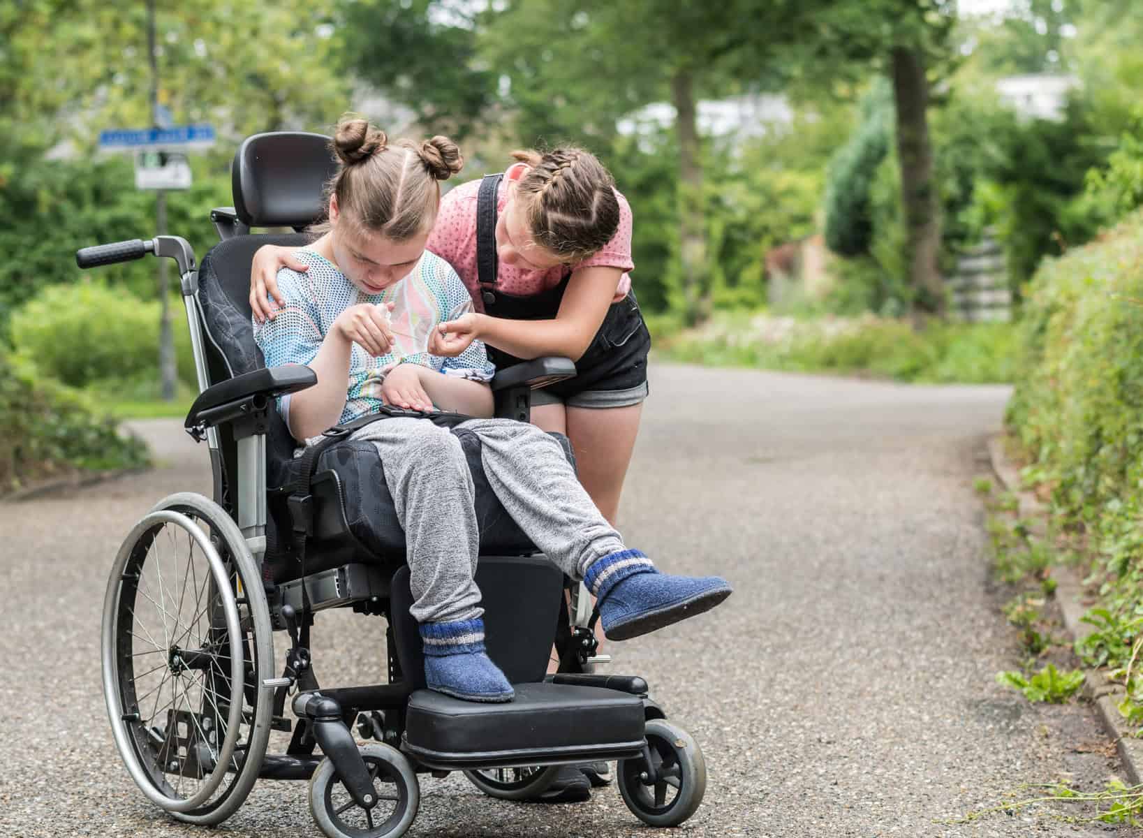 A young person in a wheelchair is being affectionately supported by another young person outdoors on a pathway, illustrating the profound impact of community support. This scene gently raises awareness about challenges similar to understanding if an iep qualification matches ssi requirements.