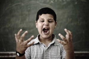 A young boy stands in front of a chalkboard, shouting with his mouth wide open and his hands raised, seeking self-regulation amid the chaos.