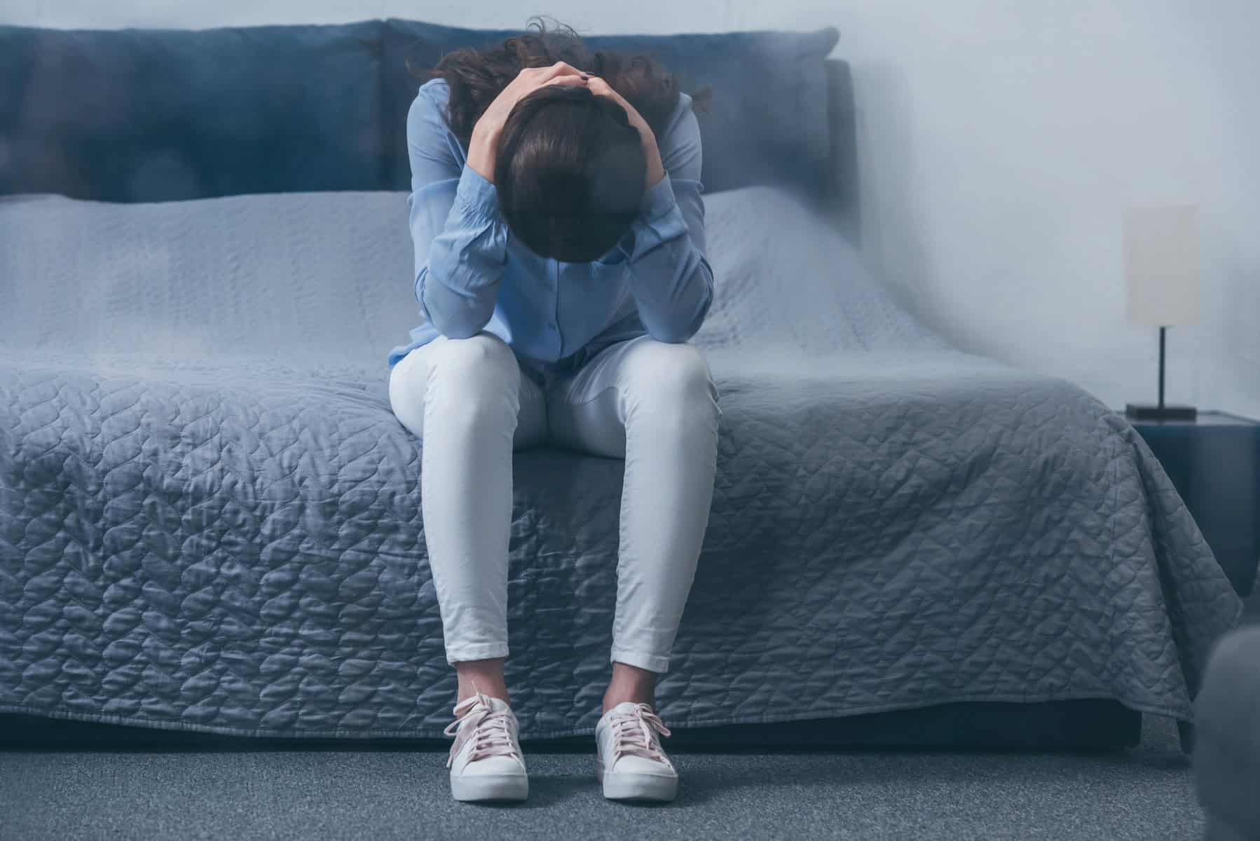 A person sits on a bed with their head in their hands, grieving their child's disability diagnosis, wearing a blue shirt and white pants.