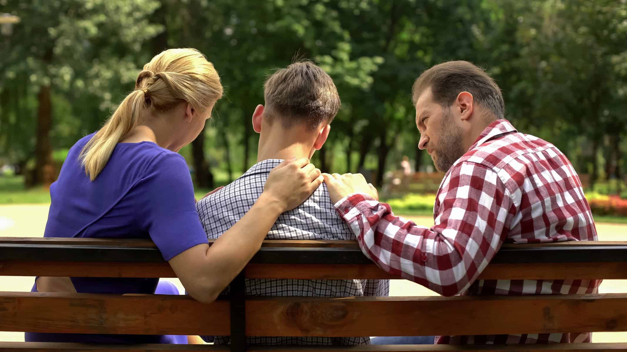 Three people sitting on a bench in the park; two place their hands on the shoulders of the person in the middle in a comforting manner, as if trying to uplift someone's spirits like an auto draft of warmth and support.