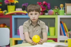 A young boy, focused on his iep goals, sits at a desk holding a yellow spiky ball, surrounded by colorful toys and books in a classroom or playroom.