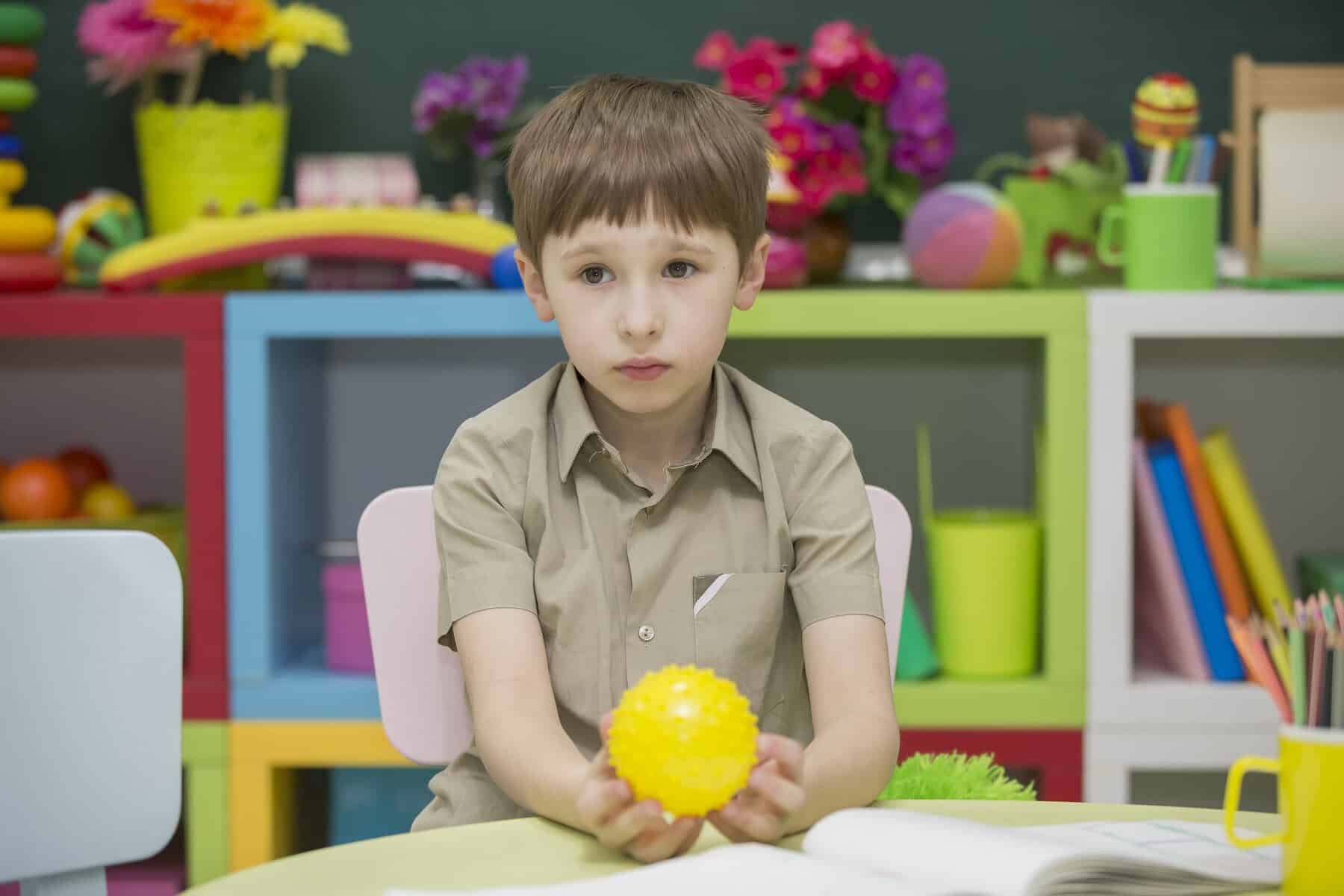 A young boy, focused on his iep goals, sits at a desk holding a yellow spiky ball, surrounded by colorful toys and books in a classroom or playroom.
