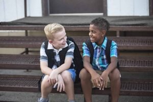 Two boys sit on metal steps, wearing backpacks and smiling at each other as they discuss conversation topics for social skills.