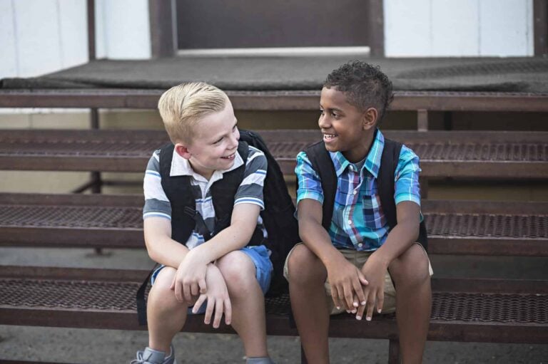Two boys sit on metal steps, wearing backpacks and smiling at each other as they discuss conversation topics for social skills.