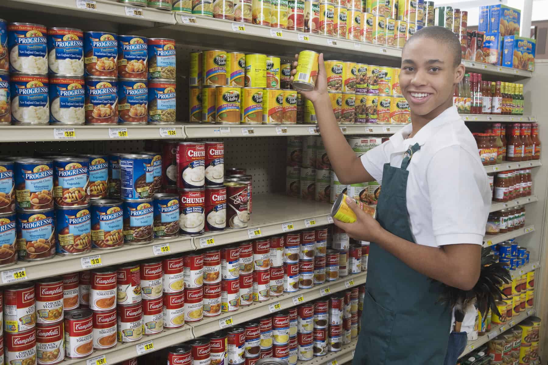 A store employee, showcasing their pre-vocational skills, organizes canned goods on shelves with precision, holding a can and smiling warmly at the camera.