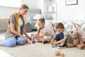 A woman and two children, one of whom is allistic, sit on a rug building with colorful blocks. A teddy bear and toy airplane are nearby.