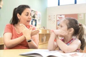 An adult woman and a young, disabled child sit at a table in a self-contained classroom, using sign language to communicate with each other over an open book.