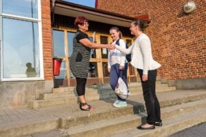 A woman greets two people outside a brick building, cultivating strong parent-teacher relationships. They are standing on concrete steps near a glass door, where effective communication is key to success.