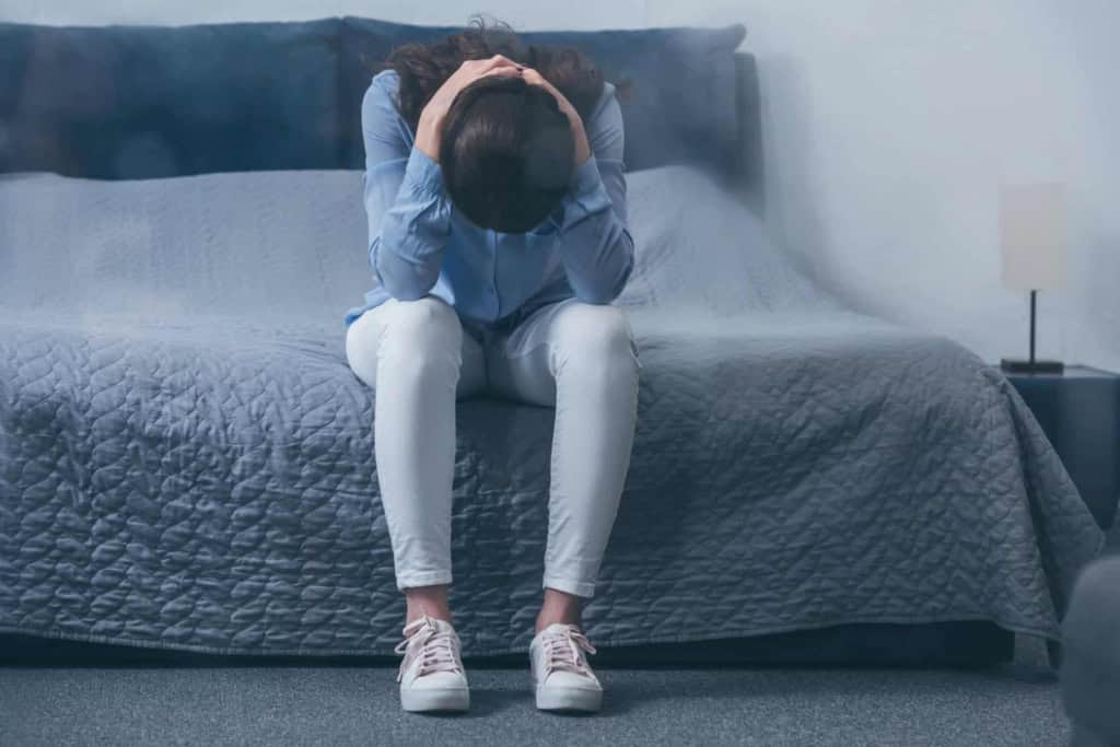 Feeling overwhelmed and stressed, woman sitting on bed with head in hands in a bedroom setting.
