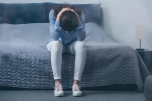 Feeling overwhelmed and stressed, woman sitting on bed with head in hands in a bedroom setting.