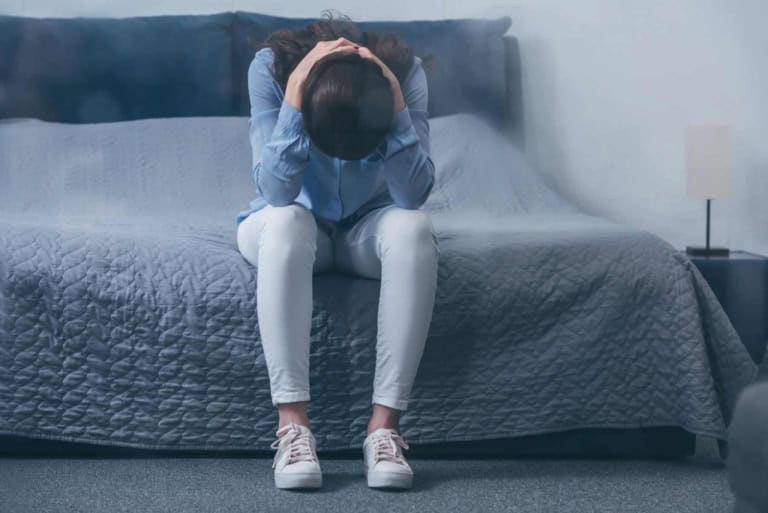 Feeling overwhelmed and stressed, woman sitting on bed with head in hands in a bedroom setting.